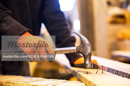 Close up of person wearing work gloves holding, hammer, removing rusty nails from recycled wooden plank.