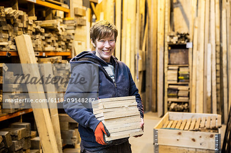 Smiling blond woman wearing work gloves standing in a workshop, holding stack of wood, looking at camera.