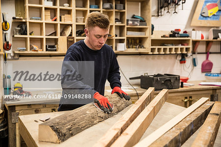 Man wearing work gloves standing at a workbench in a workshop, sanding bark of a piece of tree trunk.