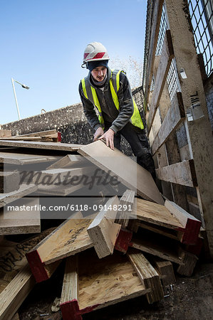 Young man wearing hard hat and reflective vest standing on top of stack of recycled wood.