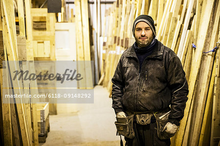 Bearded man wearing beanie and tool belt standing next to a stack of wooden planks in a warehouse, looking at camera.