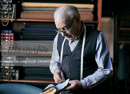 Tailor with measuring tape around his neck inspecting fabric samples.