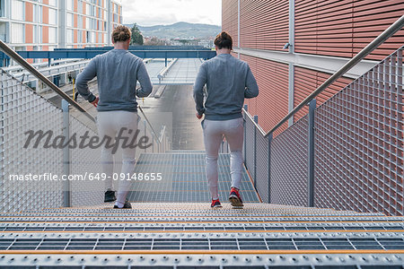Young adult male twins running together, running down city stairway, rear view