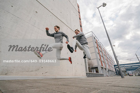 Young adult male twin runners, running and jumping mid air on city sidewalk