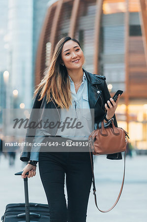 Businesswoman walking outdoors, pulling wheeled suitcase, holding smartphone and handbag, smiling