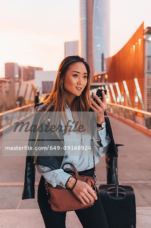 Young woman standing outdoors, using smartphone, wheeled suitcase beside her