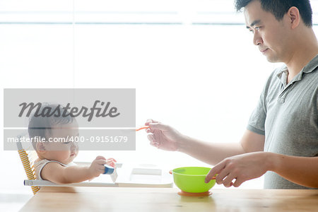 Happy Asian family at home. Father feeding solid food to his 9 months old toddler in the kitchen, living lifestyle indoors.