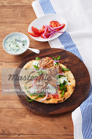 Pork souvlaki on a grilled pita on a wooden cutting board with side dishes of tzatziki and chopped tomatoes and sliced onions