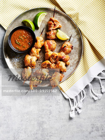 Chicken satay skewers with dipping sauce and lime wedges on a metal tray on striped fabric