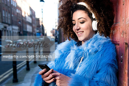 Portrait of woman with afro hair wearing fur coat, listening to music through headphones on smartphone