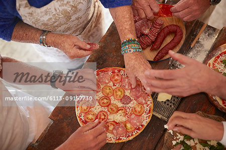 Overhead view senior friends adding tomatoes and meat to fresh pizza