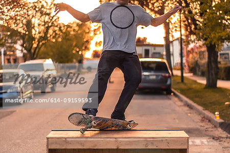 Young male skateboarder balancing on top of ramp on suburban street at sunset, neck down