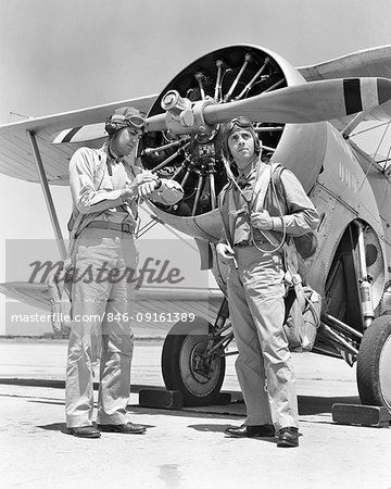 1940s TWO US NAVY PILOTS WEARING LEATHER FLYING HELMETS AND PARACHUTES STANDING READY BY PROPELLER BIPLANE