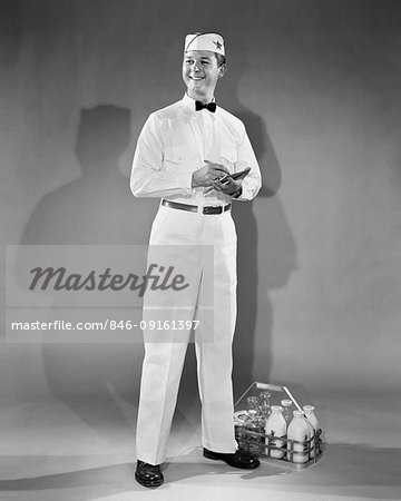 1940s 1950s SMILING HOME DELIVERY MILKMAN STANDING NEXT TO BOTTLES OF MILK HOLDING PEN AND NOTE PAD