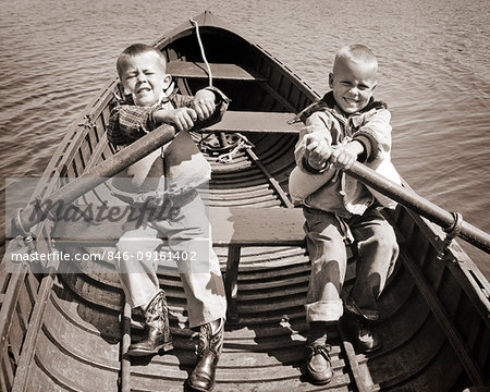 1950s 1960s PAIR OF BOYS SITTING IN BOAT EACH HOLDING ONE OAR STRUGGLING TO ROW