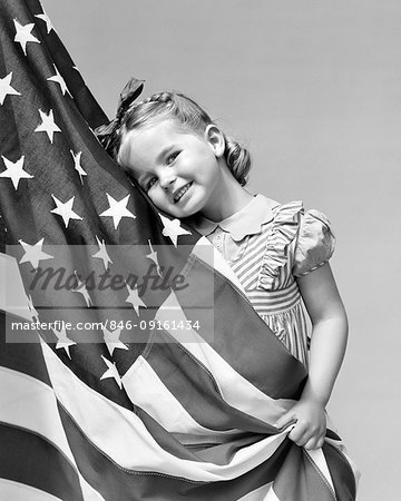 1940s LITTLE GIRL HUGGING THE AMERICAN FLAG SMILING LOOKING AT CAMERA