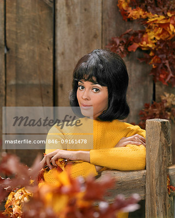 1970s AFRICAN AMERICAN YOUNG WOMAN GOLD SWEATER LEAN ON WOODEN FENCE AUTUMN LEAVES FASHION PORTRAIT LOOKING AT CAMERA