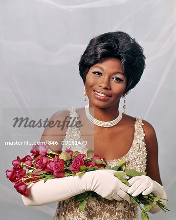 1960s 1970s SMILING AFRICAN AMERICAN WOMAN IN PEARL BEADED EVENING DRESS LONG WHITE GLOVES HOLDING RED ROSES LOOKING AT CAMERA
