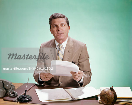 1960s BUSINESSMAN SITTING AT DESK LOOK AT CAMERA HOLDING PAPERS