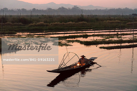 A fisherman keeps warm in his long tail fishing boat at dawn near the floating gardens on Inle Lake, Shan State, Myanmar (Burma), Asia