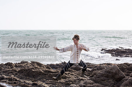 Young woman with brown hair and dreadlocks wearing white blouse standing on rocky shore by ocean, doing Tai Chi.