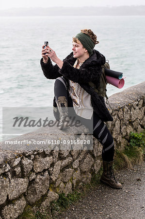 Young woman with brown hair and dreadlocks wearing headscarf sitting on dry-stone wall near ocean, taking picture with her mobile phone.