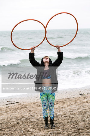 Young woman with brown hair and dreadlocks standing on a sandy beach by the ocean, balancing two hula hoops.