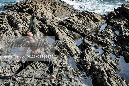 Young woman with brown hair and dreadlocks wearing sportswear standing on rocky shore by ocean, doing Tai Chi.
