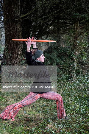 Young woman wearing floral trousers, black jacket and headscarf standing in forest, holding aloft hula hoop.