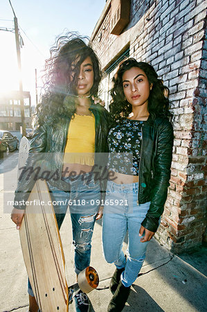 Two young smiling women with long curly black hair standing on pavement, holding skateboard, looking at camera.