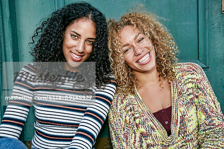 Portrait of two young women with long curly black and blond hair, smiling at camera.