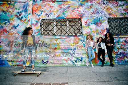 Four young women with curly hair standing in front of shutter covered in colourful graffiti, one riding a skateboard.