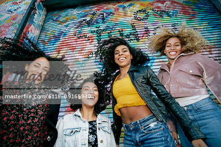 Low angle view of four young women with curly hair standing in front of shutter covered in colourful graffiti, smiling at camera.