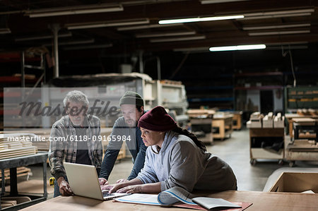 Three mixed race carpenters working on a laptop computer after work hours in a large woodworking factory.