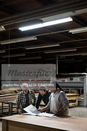 Three mixed race carpenters working on a laptop computer after work hours in a large woodworking factory.