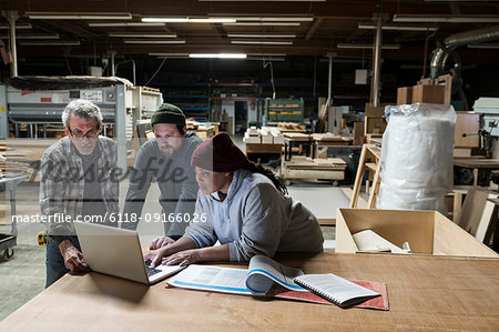 Three mixed race carpenters working on a laptop computer after work hours in a large woodworking factory.