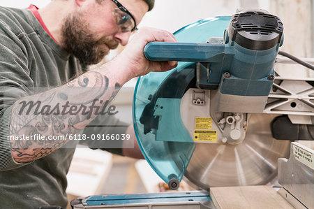 Caucasian male carpenter using a radial saw to cut a piece of wood in a large woodworking factory.