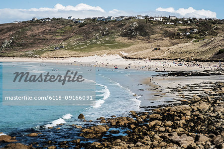 View along a coastline and sandy beach, waves breaking and people on the shore, with houses on the overlooking cliffs.