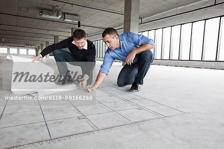 An architect and a business owner crouching over architectural plans and drawing on a concrete floor, planning a new office space in an empty raw built space.