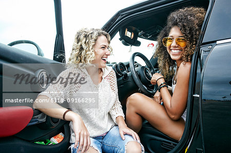 Portrait of two smiling young women with blond and brown curly hair sitting in parked car, looking at camera.