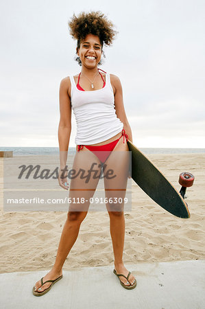 Young woman with curly brown hair wearing white vest and red bikini standing on sandy beach, holding skateboard.