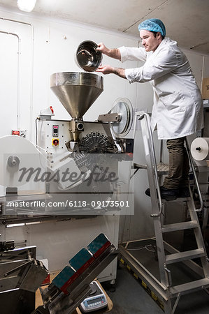 Man wearing white coat standing on a ladder, pouring tea leaves into large funnel of a machine.