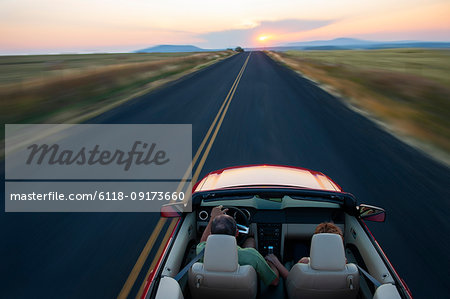 View from behind of senior couple in a convertible sports car driving on a highway at sunset in eastern Washington State, USA.