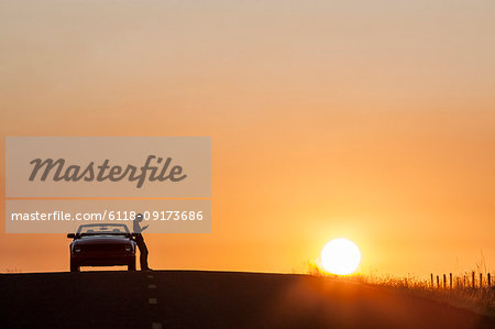 A young woman checking her phone while standing next to her car parked at a rest stop on a country road. at sunset.