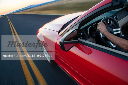 A view looking down from behind of a hand on a steering wheel of a convertible sports car moving on the highway.