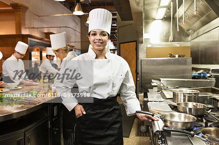 A portrait of a young black female chef in a commercial kitchen with her crew working behind her.
