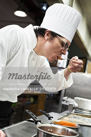 A Caucasian male chef tastes a sample of the sauce he is making in a commercial kitchen,