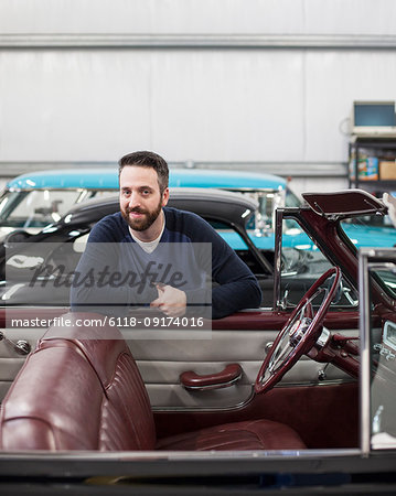 A portrait of a Caucasian male leaving on the side of his classic car convertible in a repair shop.