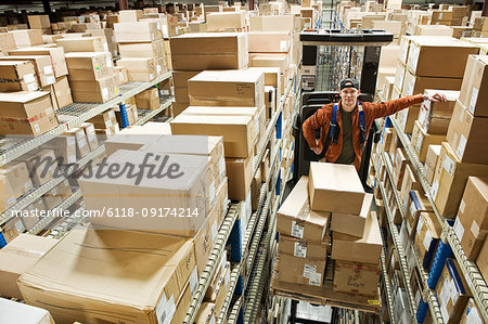 A Caucasian male warehouse worker standing on a motorized stock picker  and surrounded by products in boxes in a distribution warehouse.