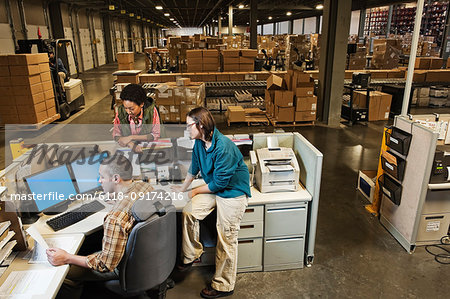 Three mixed race warehouse workers studying a document on a computer at the front desk of a distribution warehouse.